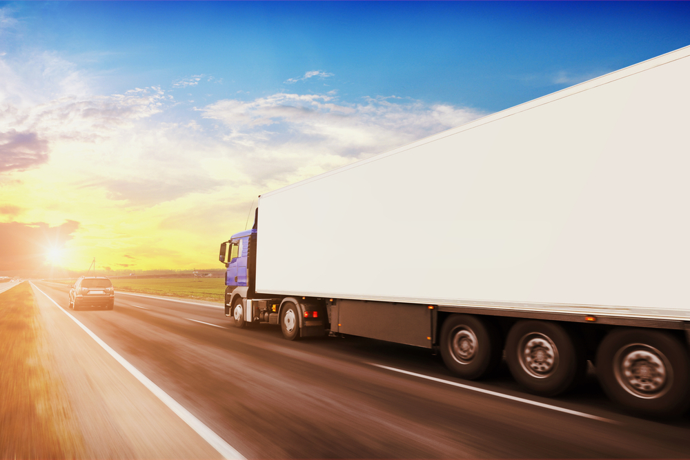 Caminhão com a frente azul e a carreta branca andando em uma por uma estrada ao por do sol